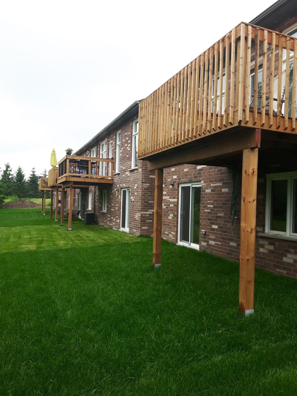 Long, elevated wood decks on a multi‑unit brick building, each supported by posts above a wide grass yard.