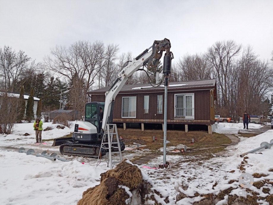 Compact excavator with a hydraulic drive installing a helical pile beside a cottage on snowy ground; a worker watches. 