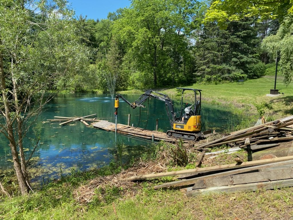 Compact excavator on a temporary timber bridge installing a helical pile at the edge of a pond in a wooded site.