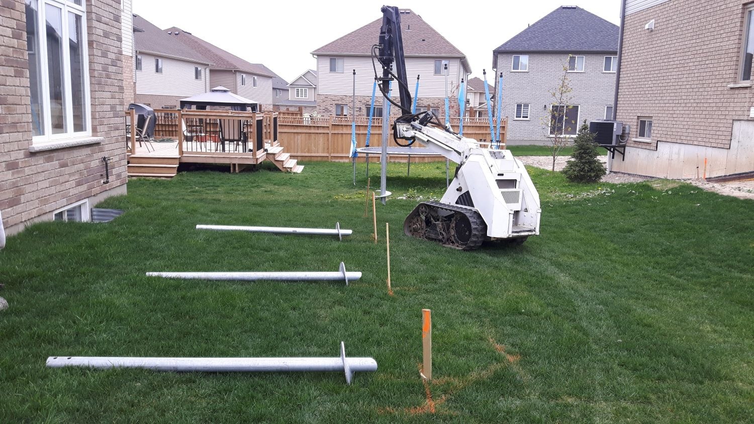 Small tracked installer with a hydraulic torque drive beside a house; steel helical piles staged on the backyard lawn.