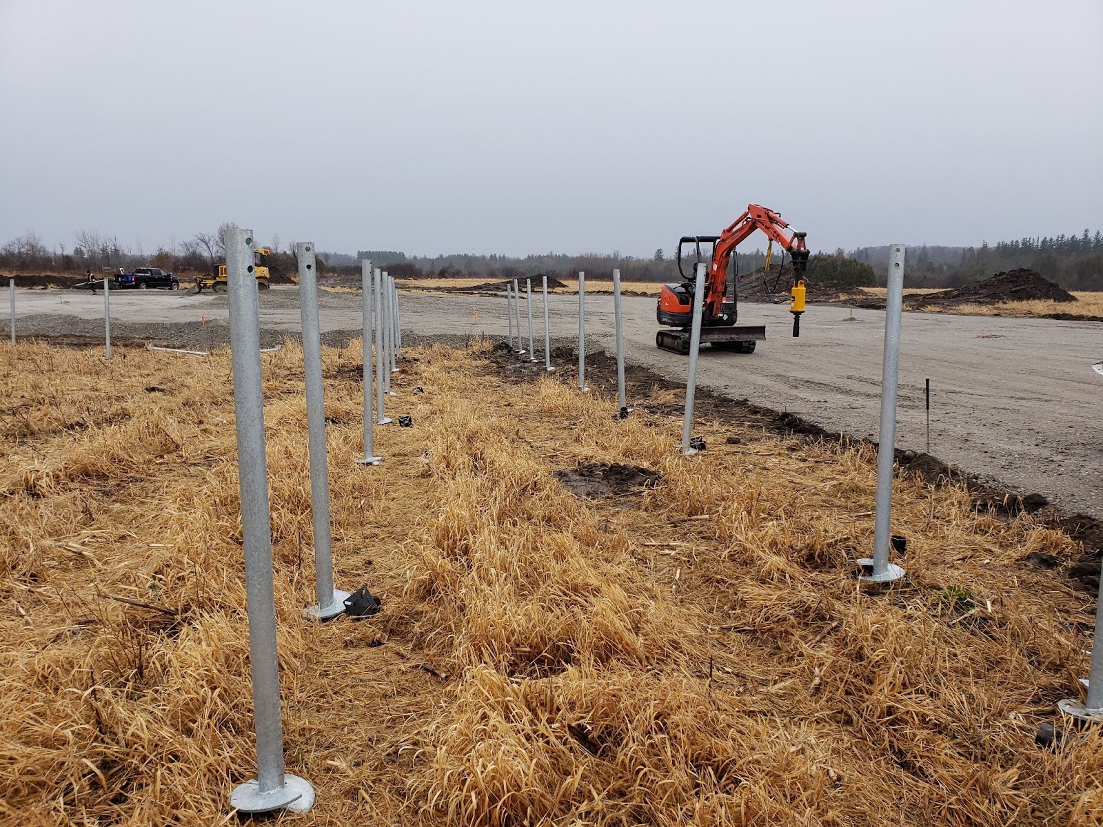 Row of newly installed galvanized helical piles across a field; excavator and worker setting additional piles in the background.