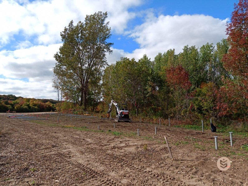 Excavator working from a timber causeway across a pond to install helical piles at a green, wooded site.