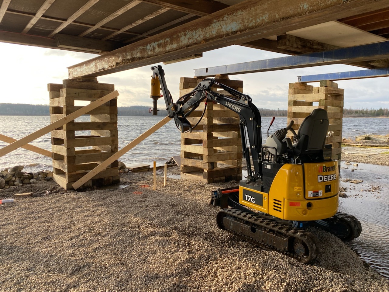 Compact excavator working under stacked timber cribbing used as a reaction frame during a helical pile test.