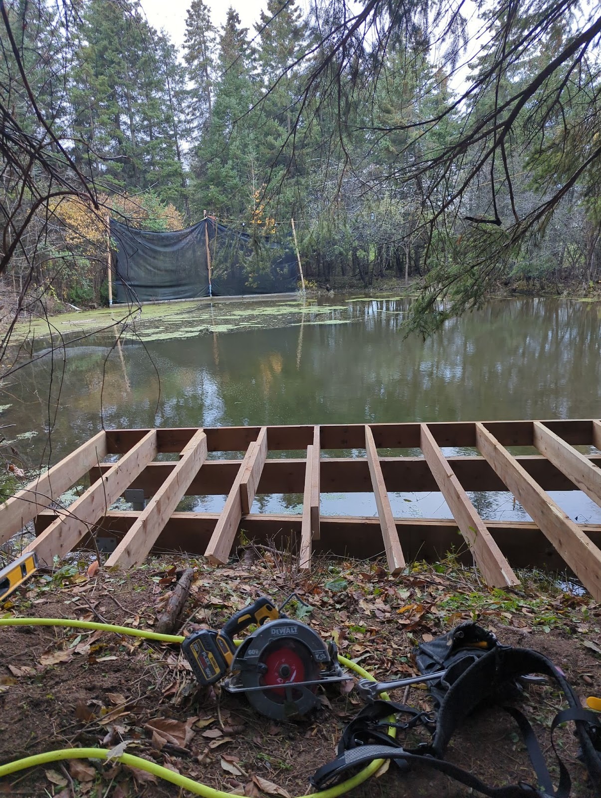 Deck joist frame under construction at the edge of a pond; circular saw and hoses in the foreground.