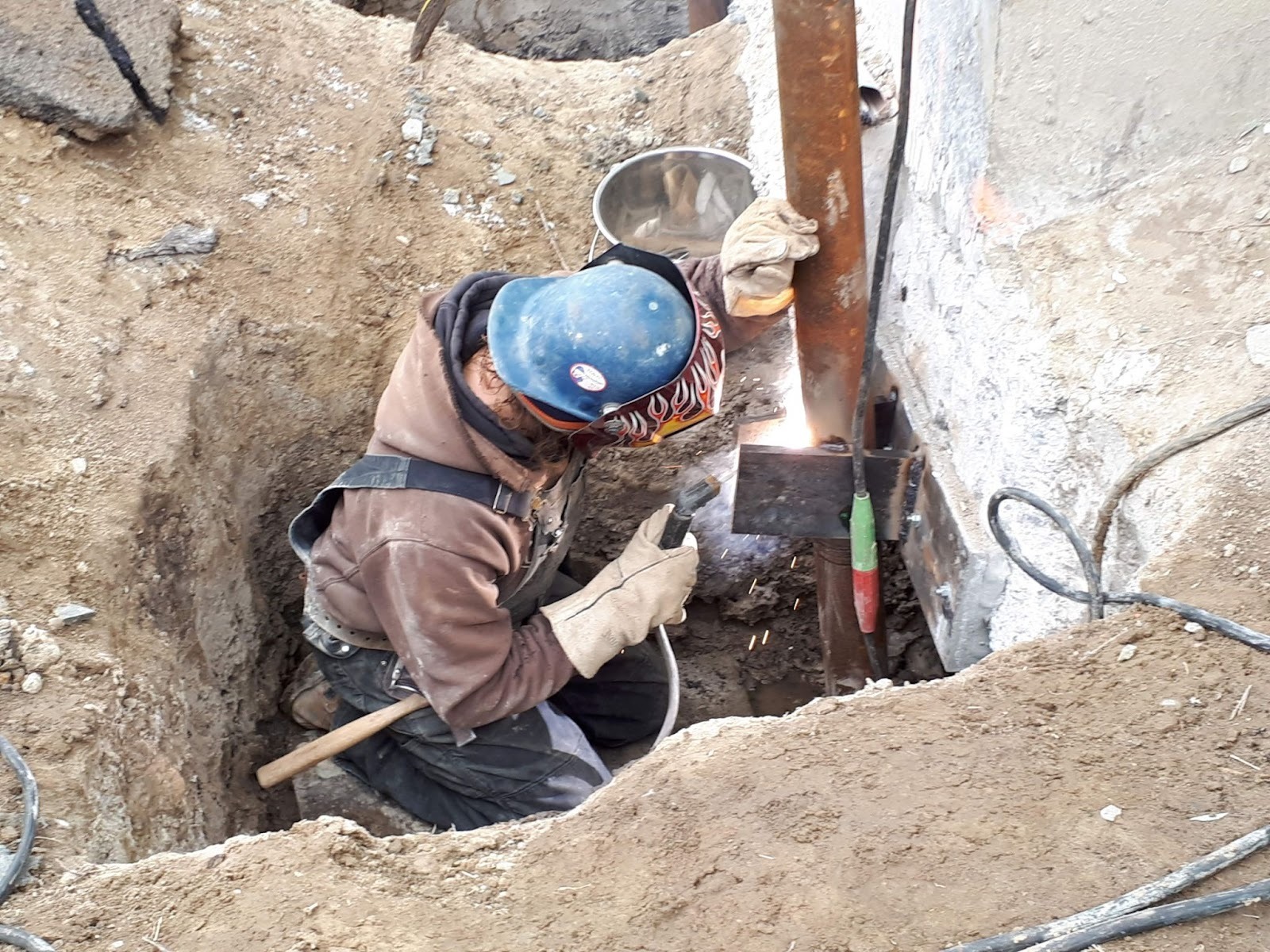 Worker in a trench operating a jack on a steel pile near a foundation wall; hoses and cables connected.