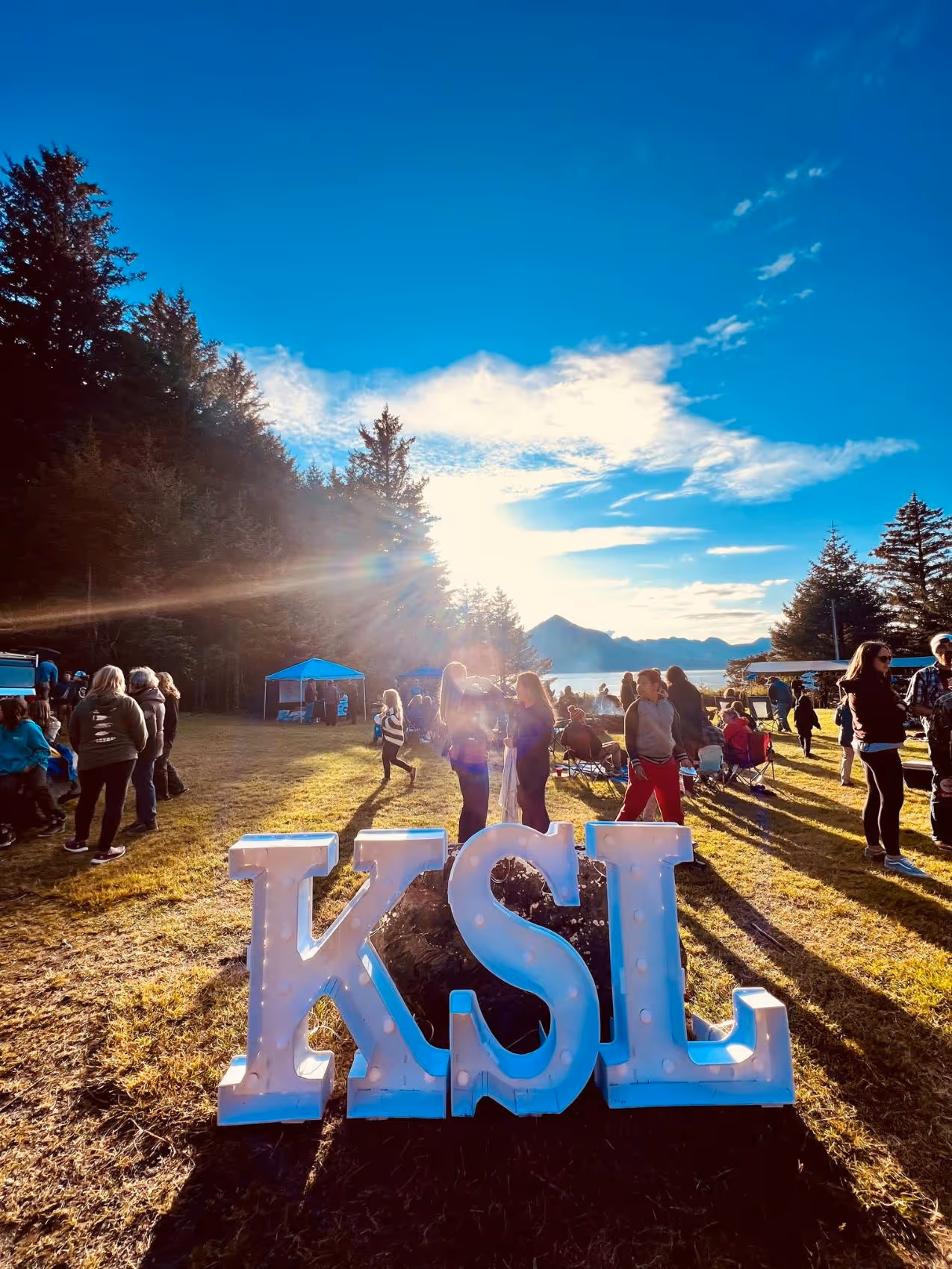 Outdoor gathering of people at sunset near large illuminated letters spelling KSL on grass, surrounded by trees and mountains in the background.