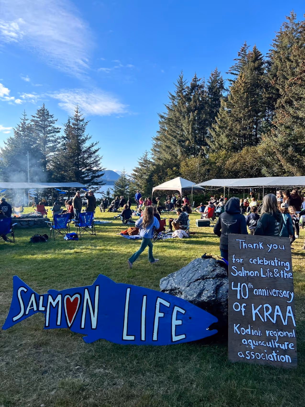 Outdoor gathering with people sitting on lawn chairs and a sign shaped like a blue fish reading 'SALMON LIFE' alongside a board thanking for the 40th anniversary of Kodiak Regional Aquaculture Association.