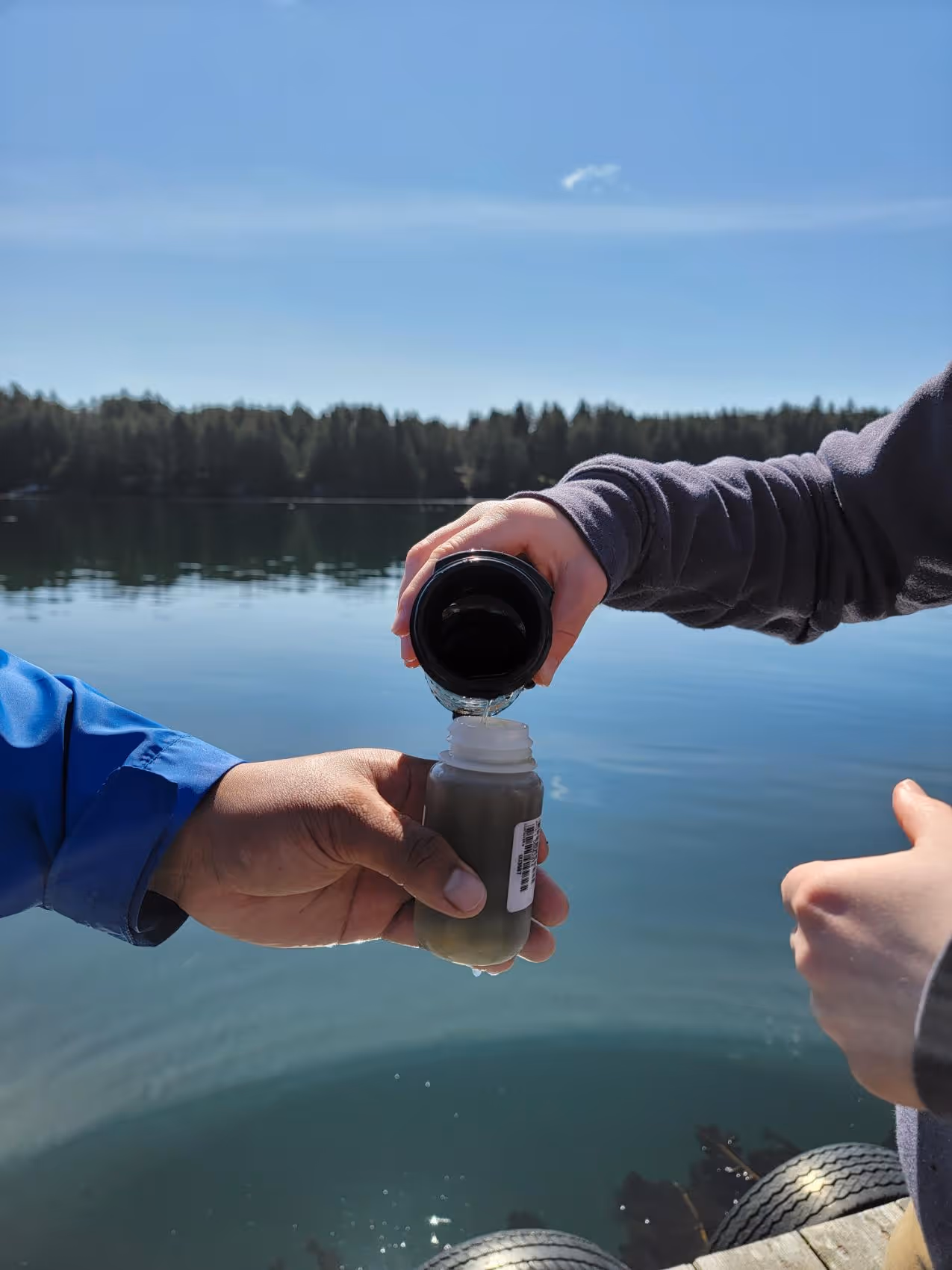 Two hands near a lake, one pouring water from a black container into a small bottle held by the other hand.