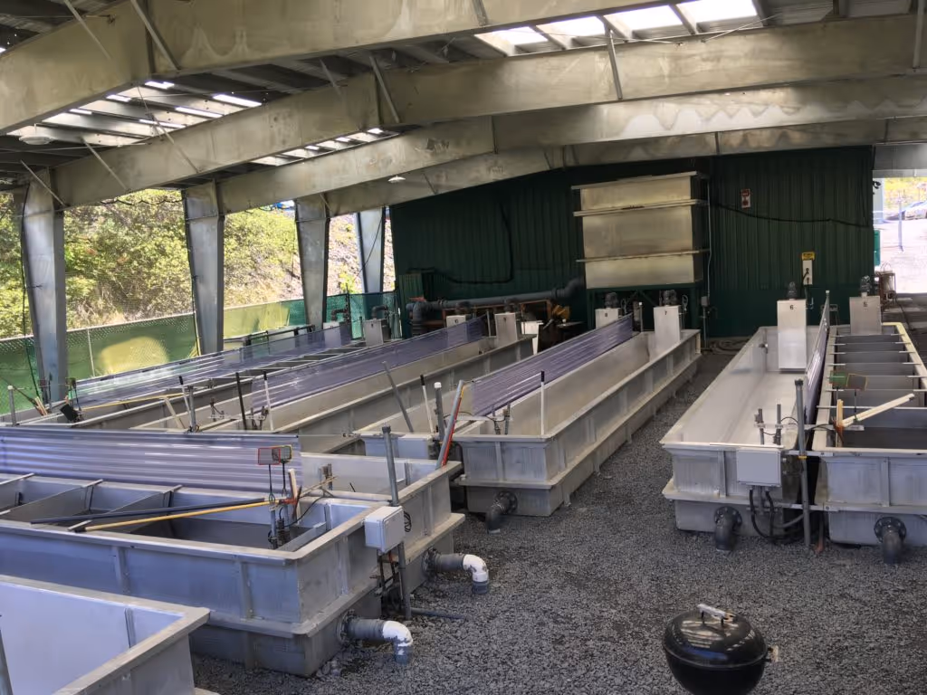 Industrial indoor facility with large rectangular water tanks and metal pipes on a gravel floor under a partially open roof.