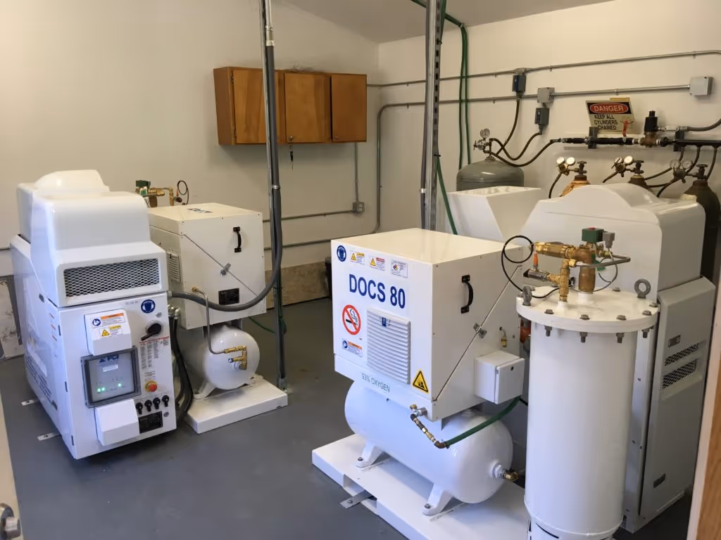 Industrial room with white medical oxygen concentrators including one labeled DOCS 80 and gas cylinders in the background.