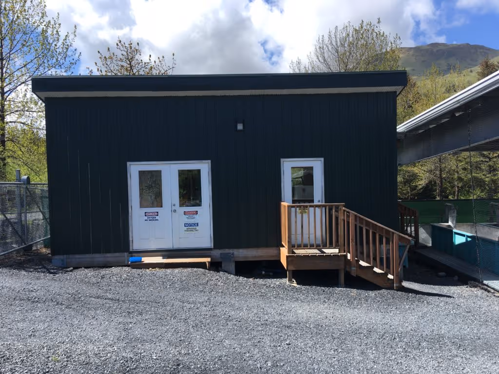 Small dark green building with two white doors, one with a wooden ramp and railing, situated on gravel with trees and mountains in the background.