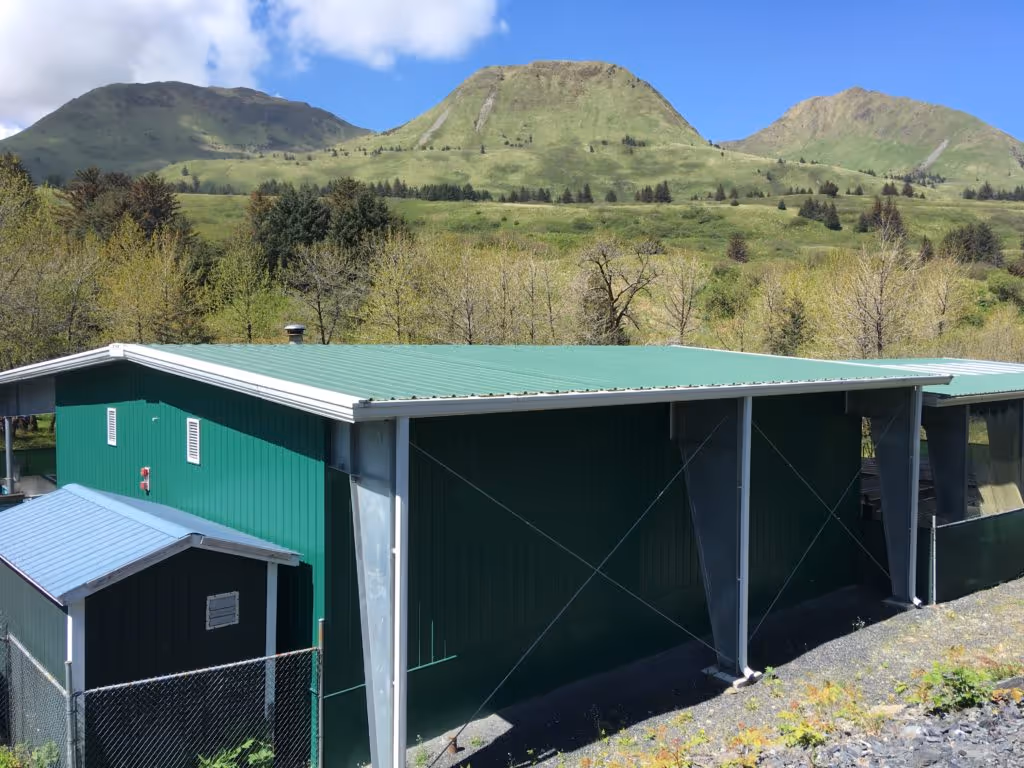 Green metal building with a shed roof surrounded by trees and hills under a blue sky with clouds.