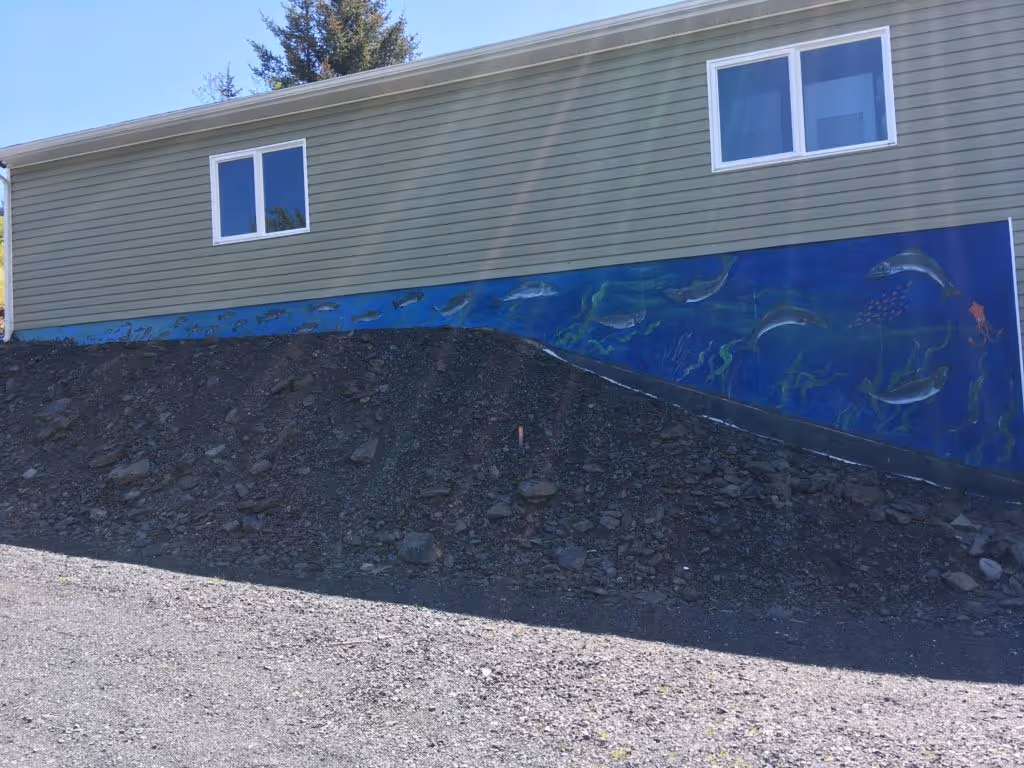 Side of a green house with two windows and a mural at the base depicting underwater sea life, including fish, dolphins, seaweed, and an octopus against a blue background.