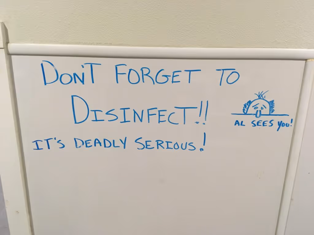 Whiteboard with message in blue marker reading 'DON'T FORGET TO DISINFECT!! IT'S DEADLY SERIOUS!' and a drawing of a face peeking over a line with text 'AL SEES you!'