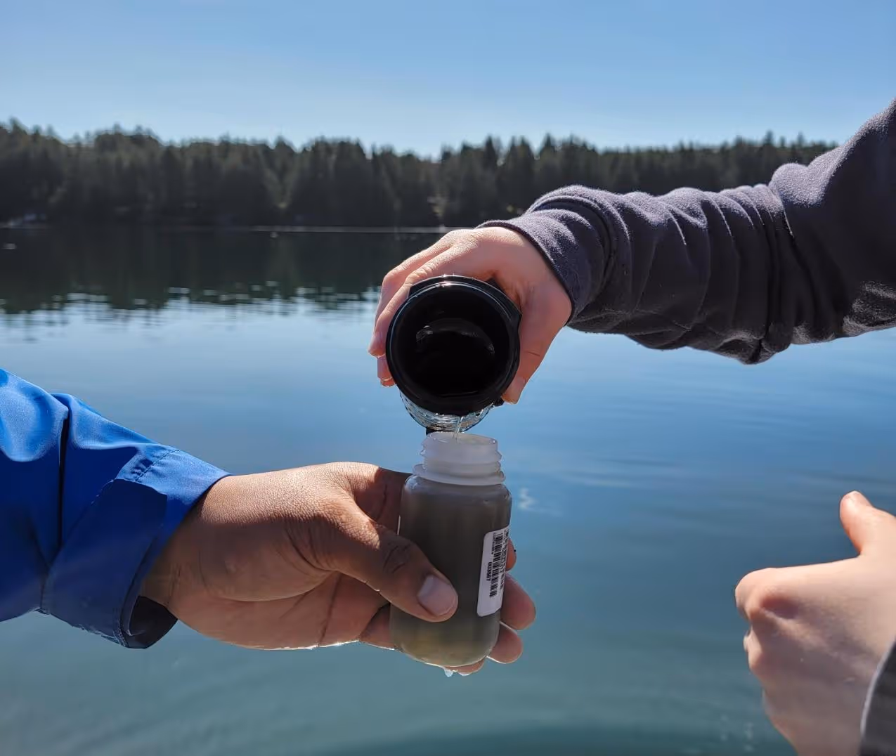 Two hands near a lake, one pouring clear water from a black bottle into a small transparent container with a barcode label.