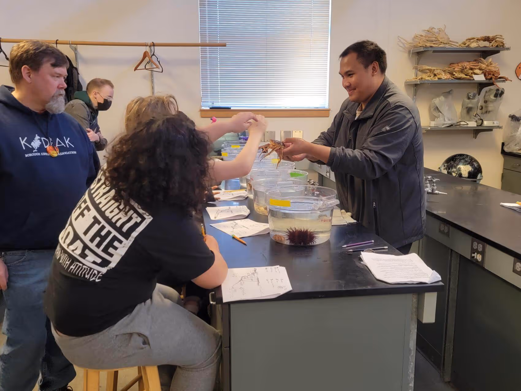 A group of people in a classroom examining marine animals in containers, with one person holding a crab and others observing or taking notes.