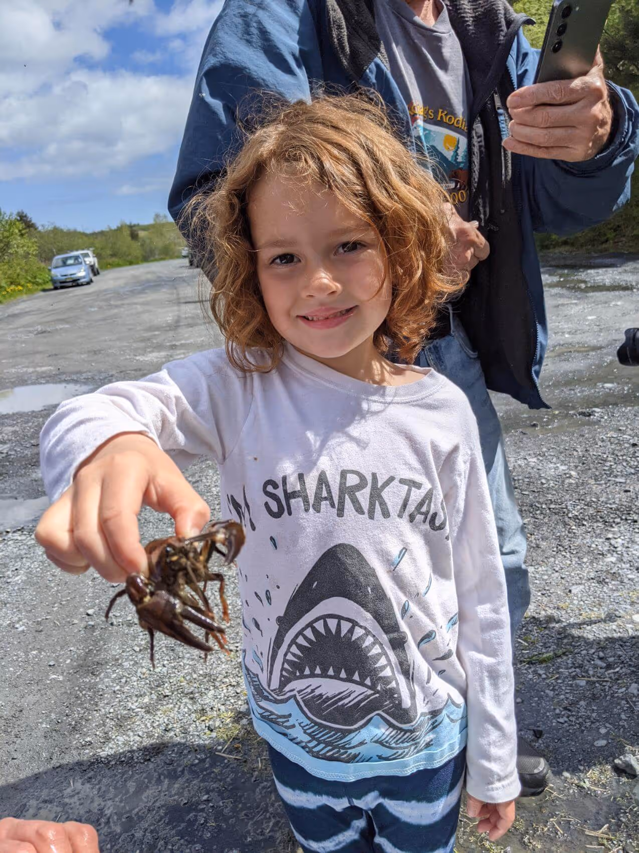 Child with curly hair wearing a white shirt with a shark graphic, holding a small crab outdoors on a gravel path.