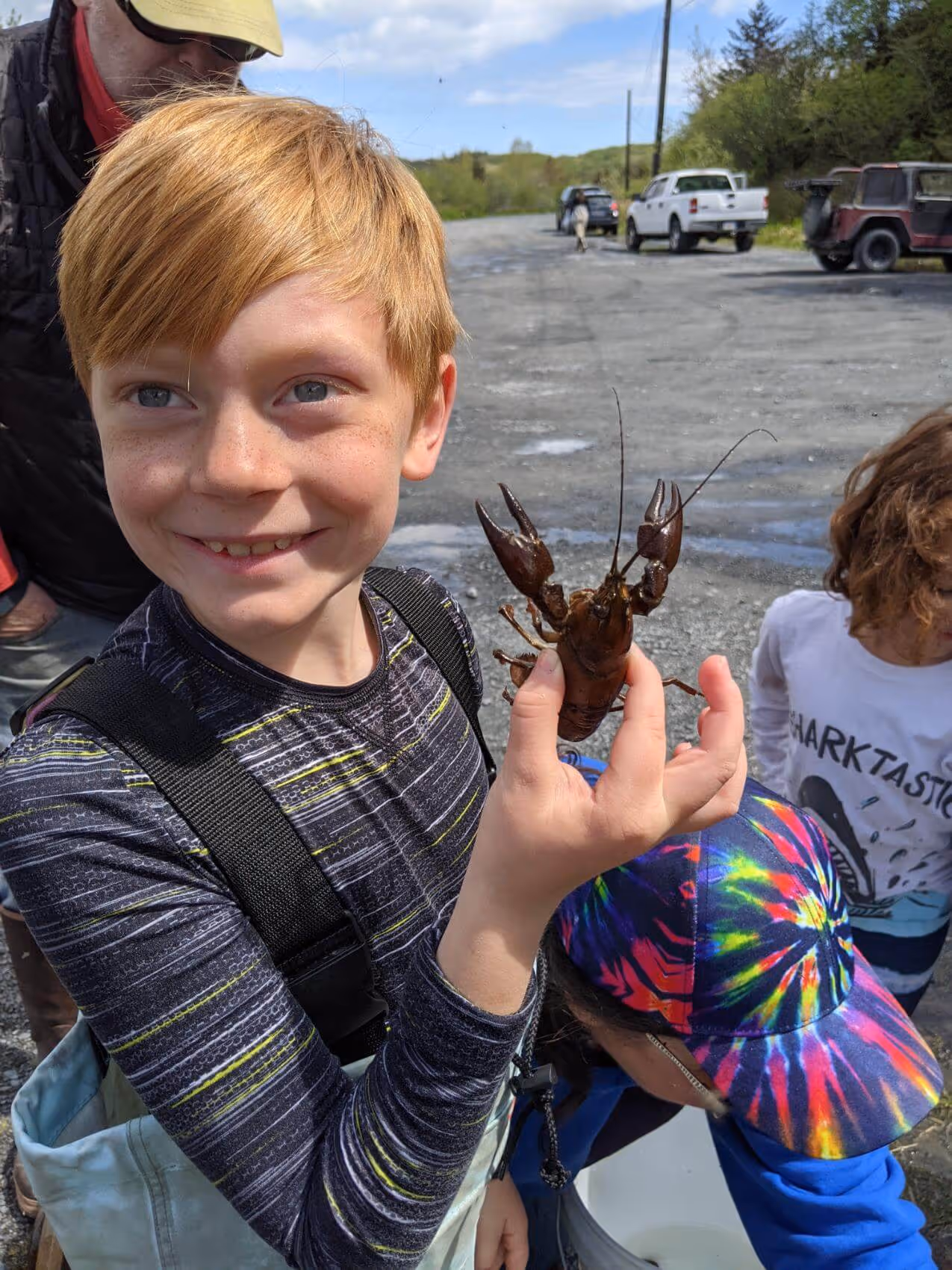 Smiling red-haired boy holding up a crawfish outdoors with other children and vehicles in the background.