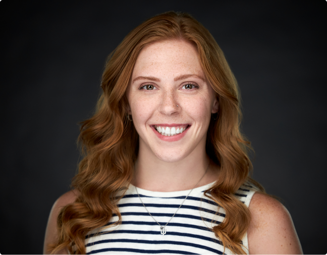 Smiling woman with long wavy red hair, freckles, wearing a striped sleeveless top and a necklace.