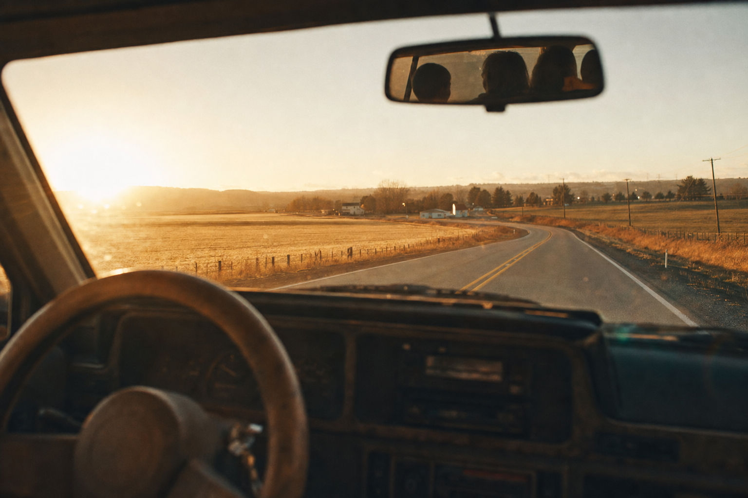 View through a car windshield of a two-lane rural highway at golden hour, with a worn steering wheel and dashboard in the foreground.