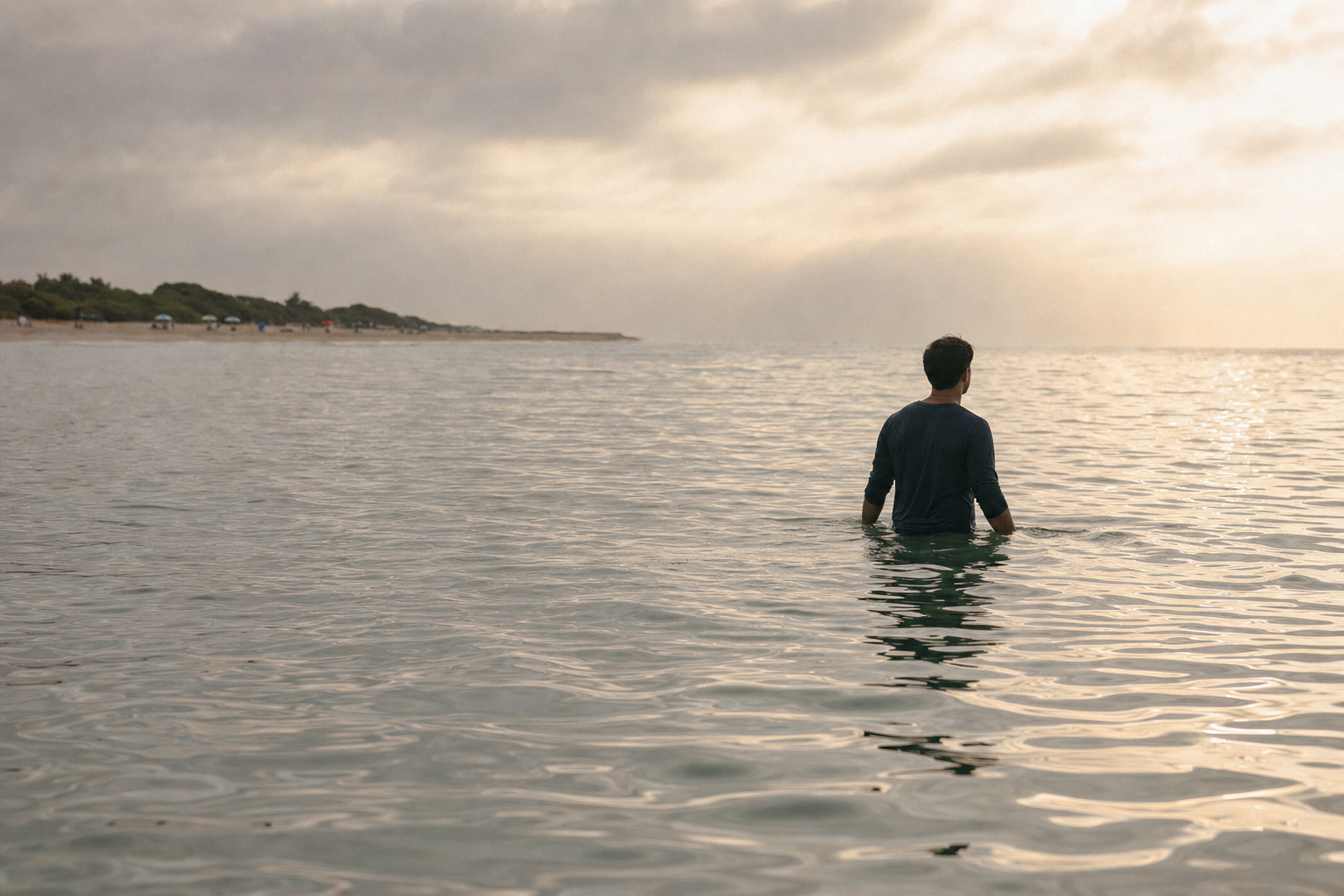 A lone person standing waist-deep in calm ocean water, seen from behind, with a distant shoreline and beach umbrella far behind them.