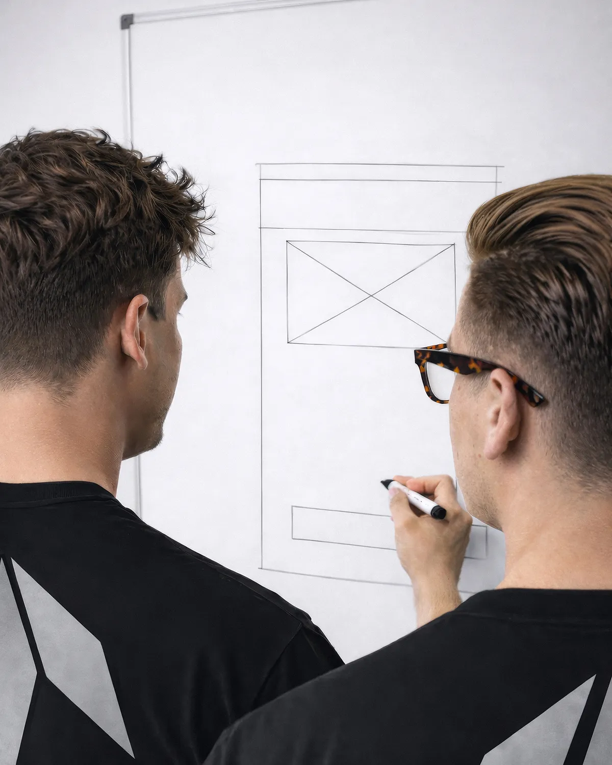 Dwóch mężczyzn w czarnych koszulkach rysuje projekt strony internetowej na białej tablicy.