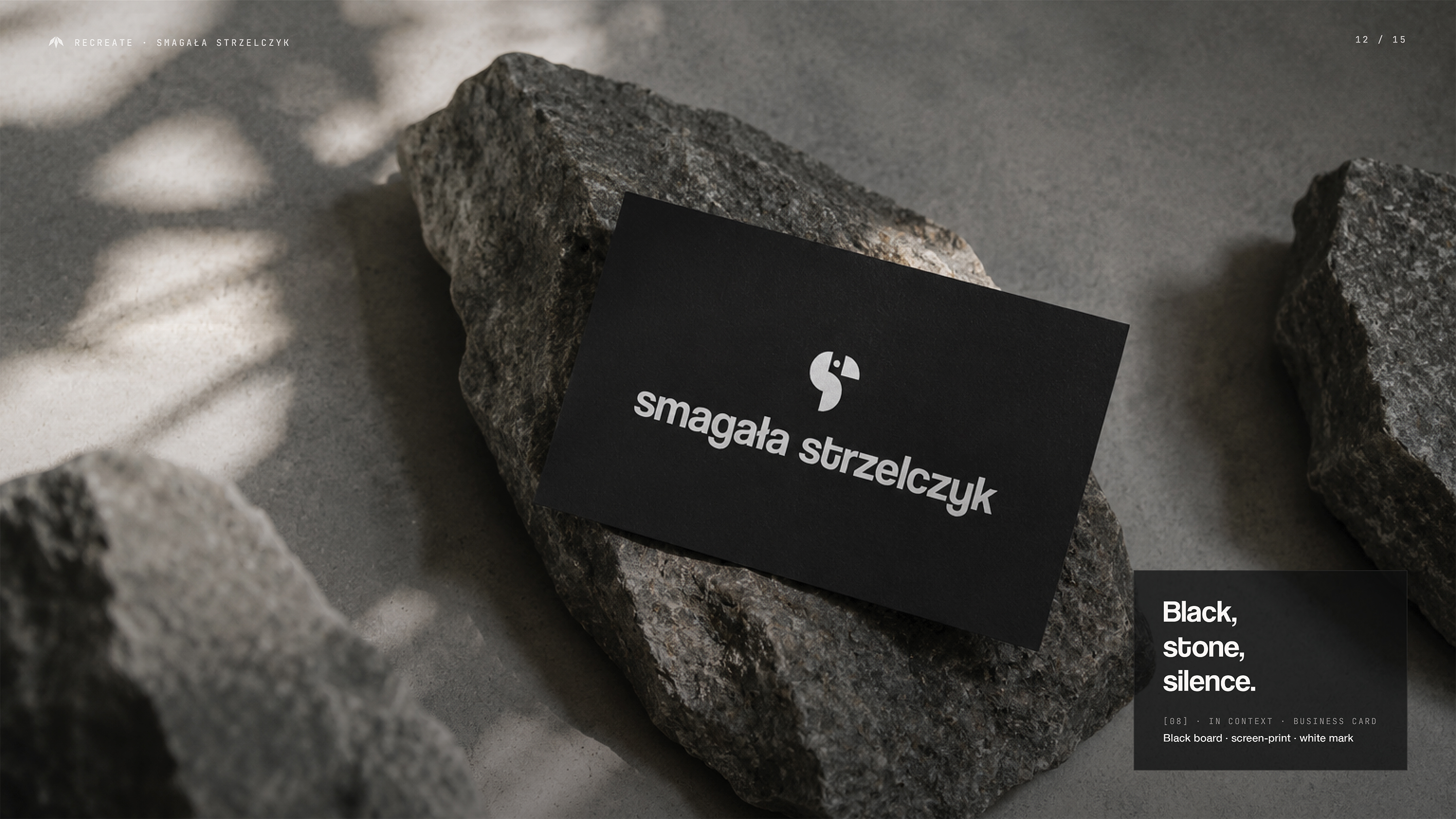 Czarna wizytówka Smagała Strzelczyk z białym logo papugi i wordmarkiem leży na szorstkiej granitowej skale — boczne naturalne światło rzuca cień liści; opis: Black, stone, silence; screen-print white mark on black board