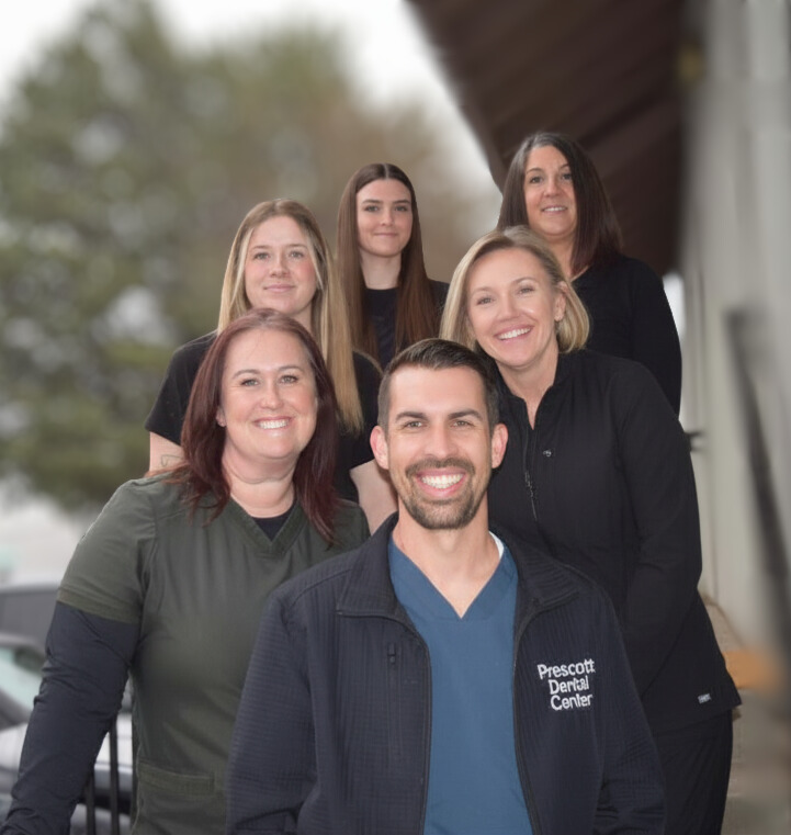 Group portrait of six smiling dental professionals standing outdoors, with the man in front wearing a jacket labeled Prescott Dental Center.
