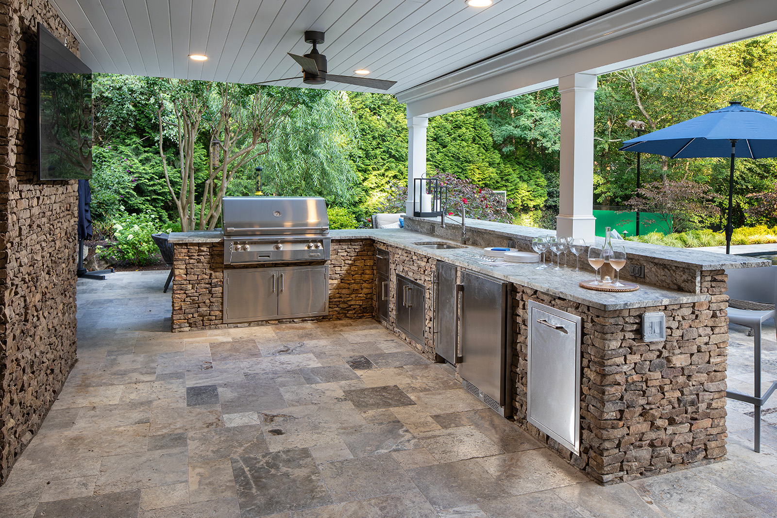 A beautiful stone outdoor kitchen with grill, wine glasses, and TV, built by Rossen Landscape.