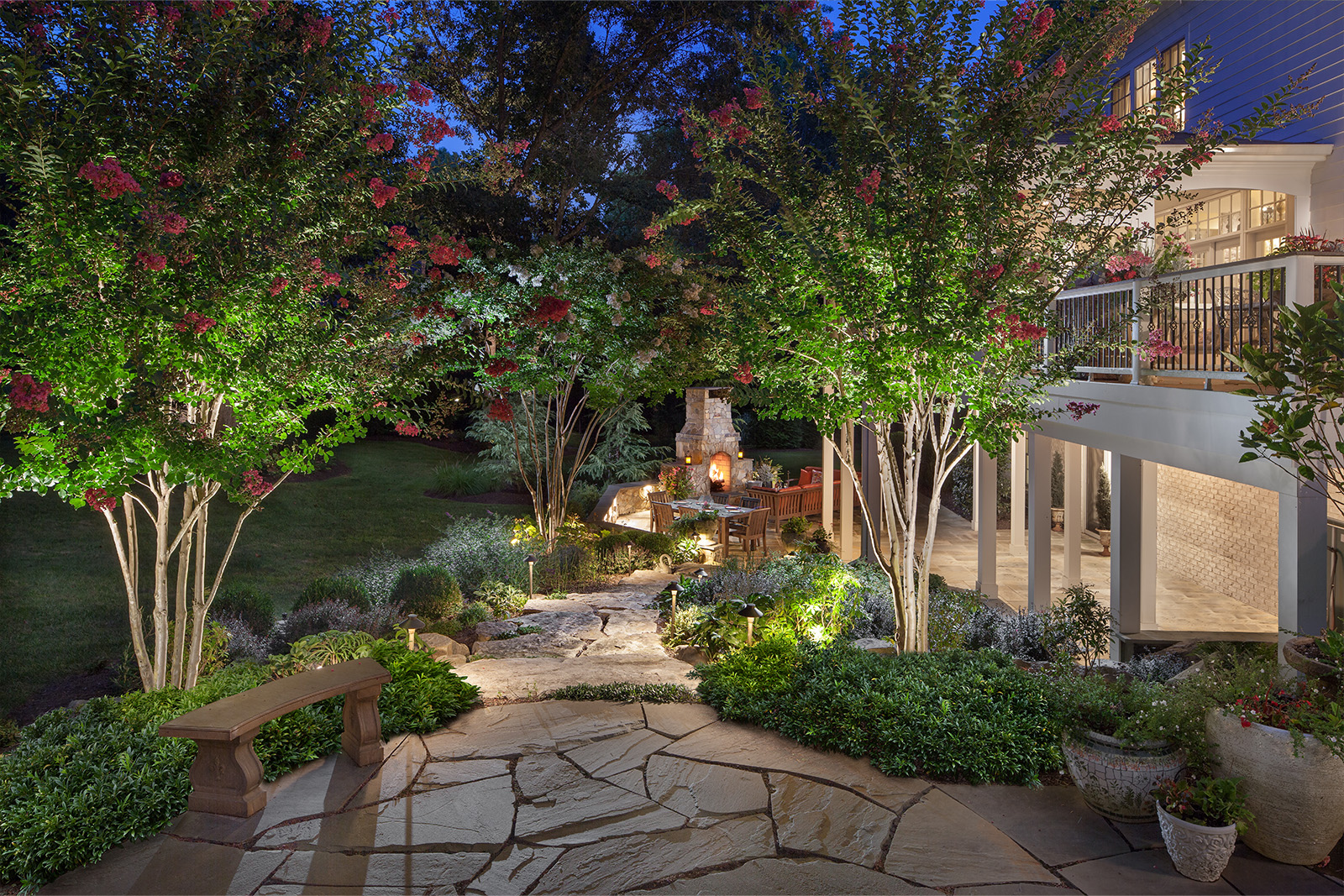 A beautiful backyard landscape with shrubs, flowers, and tree surrounding a seating area with a stone fire place. The area is well lit with lights.