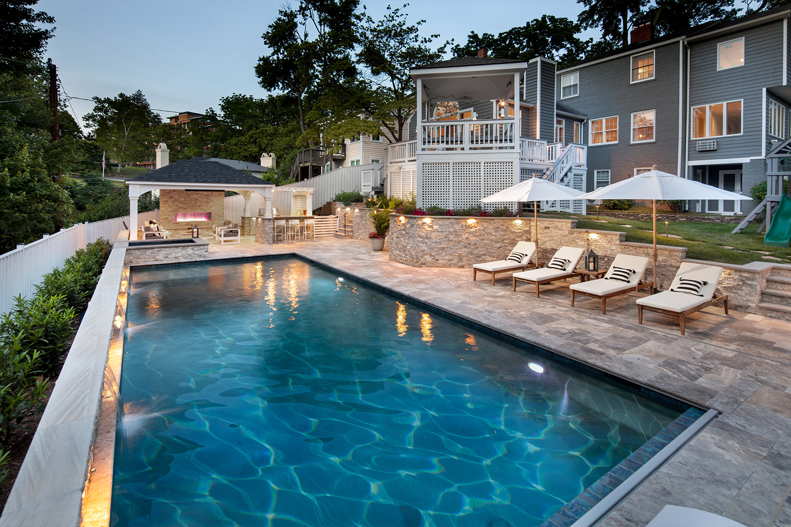 A stunning backyard pool with lounge hairs, an outdoor kitchen, and beautiful lighting illuminating the area.