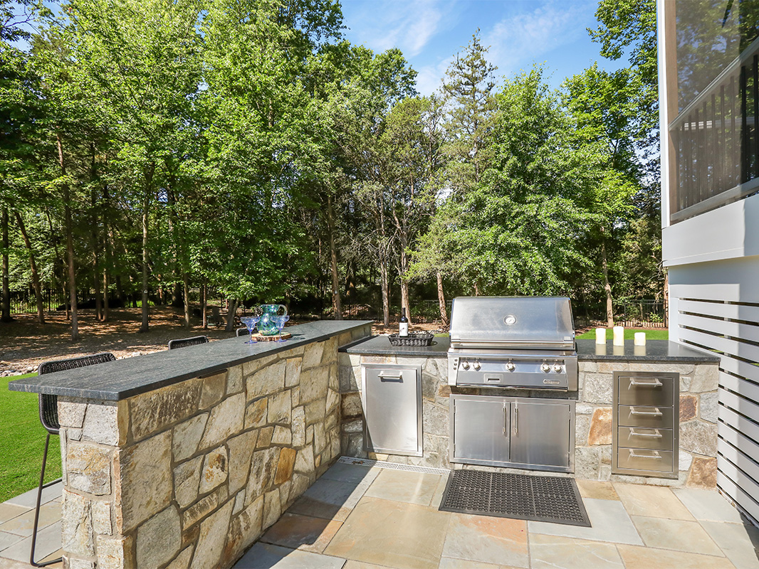 Gorgeous backyard retreat with stone patio, lounge chairs, brick wall, outdoor grill station, and luscious grass. This project was designed and built by Rossen Landscape.