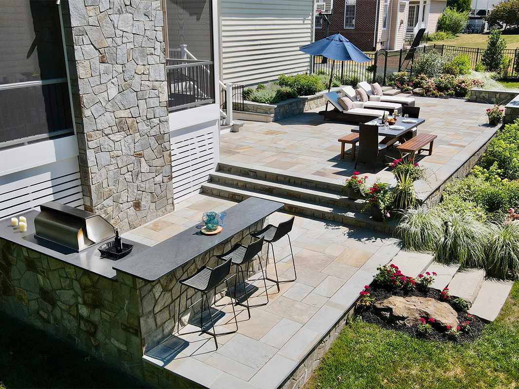 Gorgeous backyard retreat with stone patio, lounge chairs, brick wall, outdoor grill station, and luscious grass. This project was designed and built by Rossen Landscape.