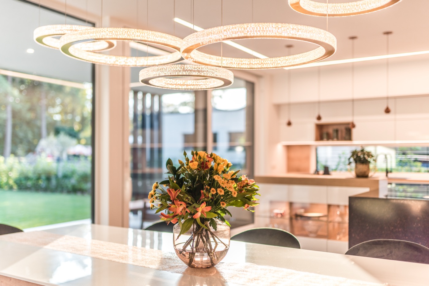 Nahaufnahme eines Esstisches mit einem farbenfrohen Blumenstrauß, über dem mehrere ringförmige, funkelnde Designer-Leuchten hängen