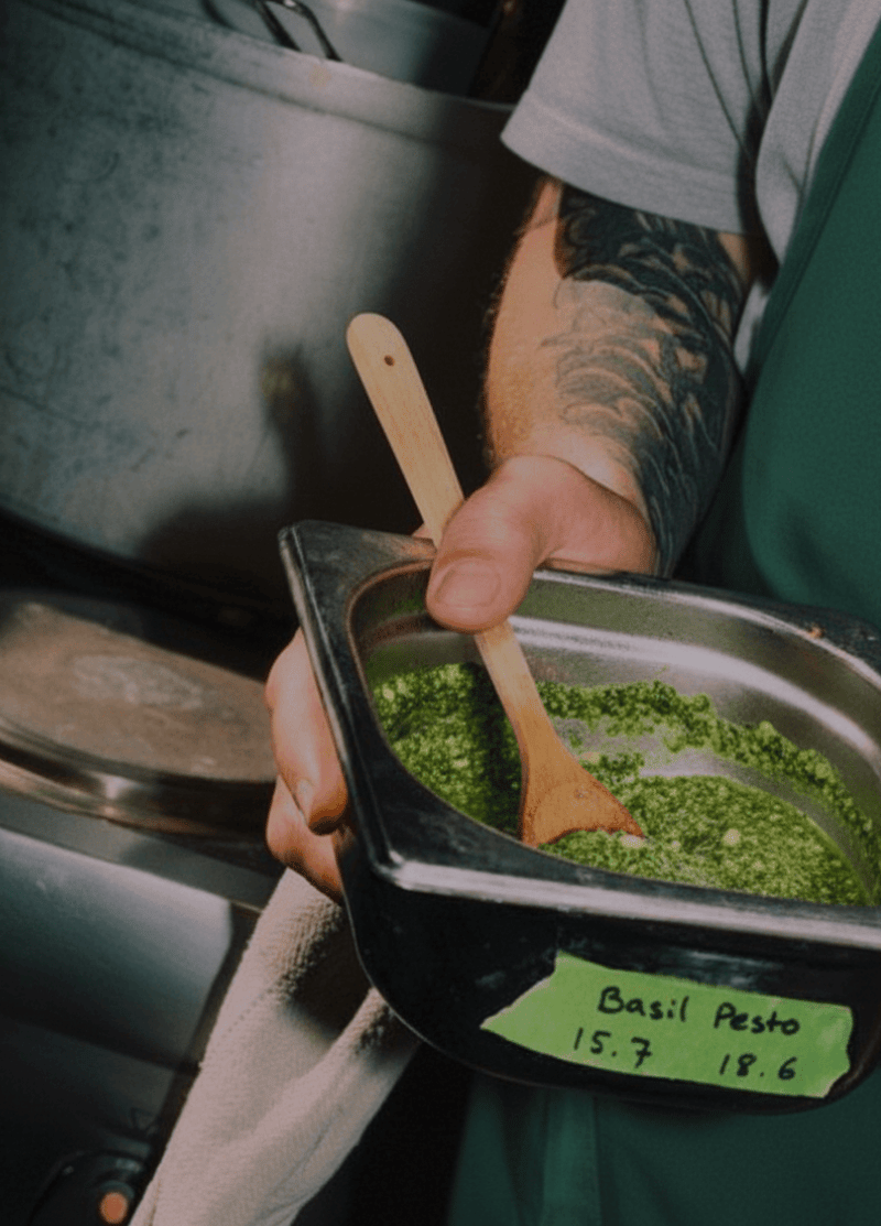 Person with tattooed arm holding a container of green basil pesto with a wooden spoon in a kitchen setting.