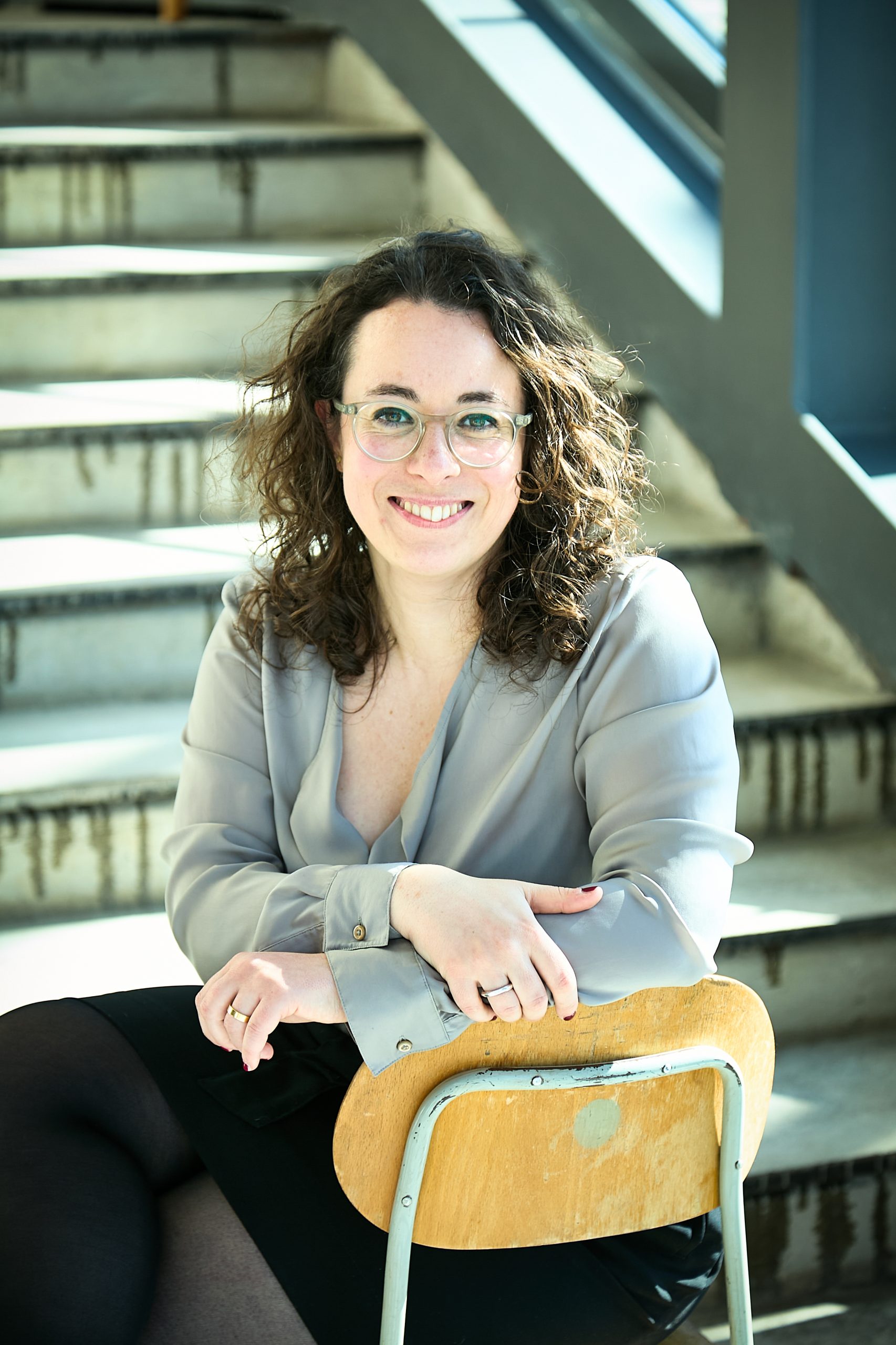 Smiling woman with curly hair and glasses sitting on a wooden chair in front of concrete stairs with sunlight.