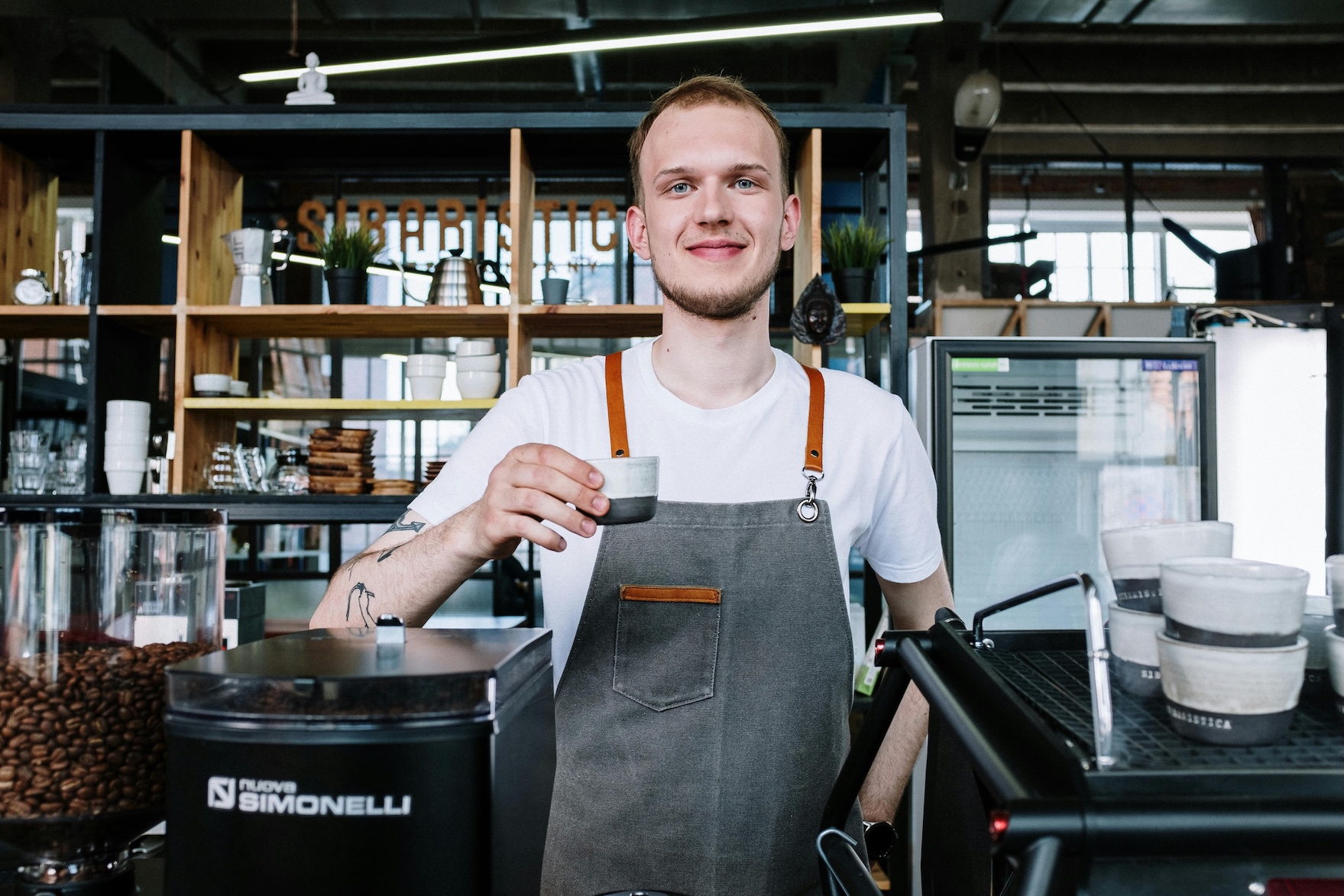 Barista in gray apron holding a small coffee cup behind a coffee machine in a café.