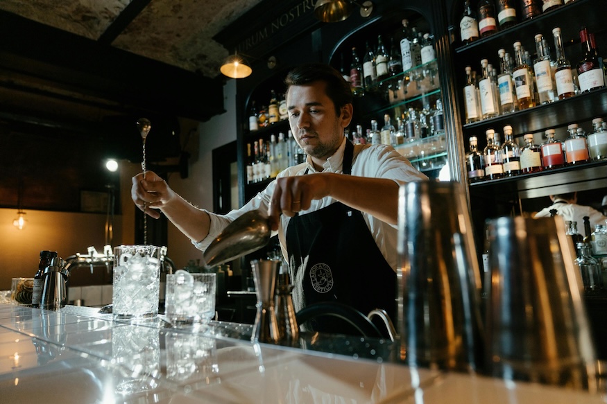 Bartender in a black apron pouring a drink at a bar with shelves of liquor bottles in the background.