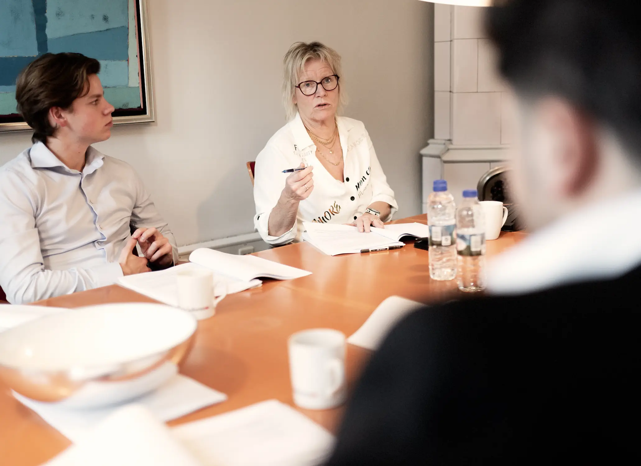 En kvinde med briller sidder ved et mødebord og taler, mens hun holder en pen. Ved siden af hende sidder en mand i skjorte og lytter. På bordet ligger åbne notesbøger og papirer, og der står vandflasker og kopper.