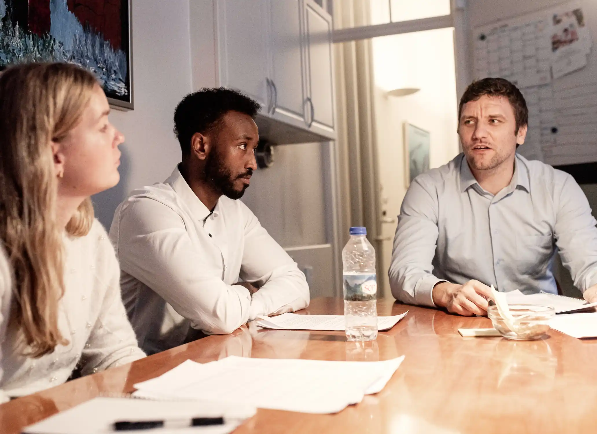 Tre kolleger sidder omkring et mødebord på et kontor og taler sammen. En mand i hvid skjorte lytter med armene over kors, mens en mand i lyseblå skjorte ser ud til at forklare noget. Der ligger papirer på bordet, og en vandflaske står i midten.