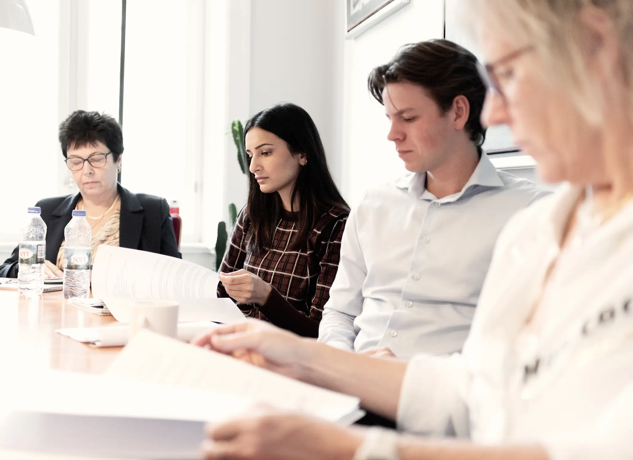 Fire personer sidder ved et mødebord i et lyst kontor og læser i dokumenter. En kvinde i ternet bluse gennemgår et papir, mens en mand i lys skjorte sidder ved siden af og kigger ned. I forgrunden ses en person ude af fokus med papirer, og på bordet står vandflasker og kopper.