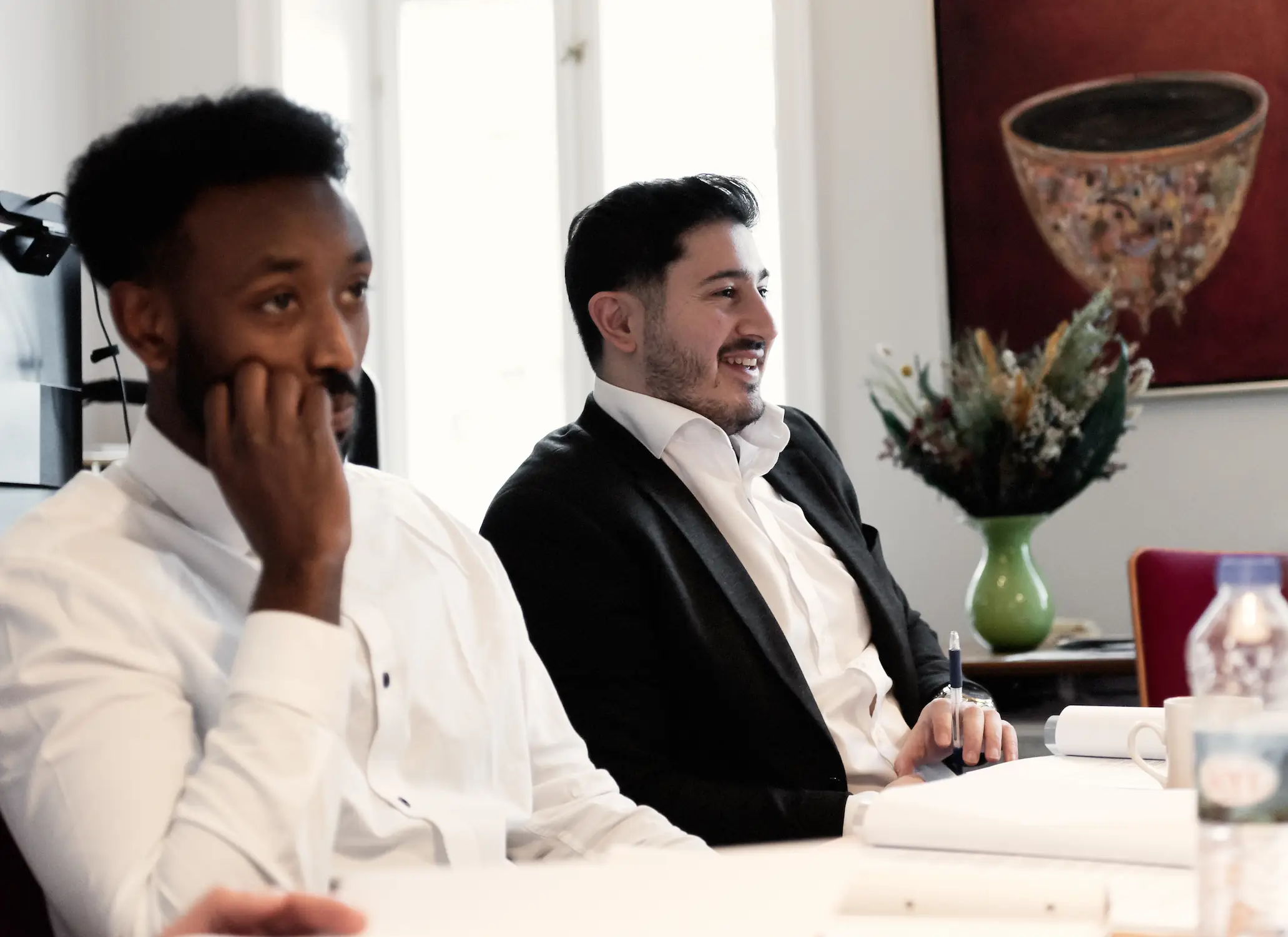 To mænd sidder ved et mødebord i et lyst kontor under et møde. Den ene læner sig tilbage og lytter med hånden ved kinden, mens den anden i jakkesæt taler og holder en pen. På bordet ligger mapper og papirer, og i baggrunden ses en vase med blomster og et vægkunstværk.