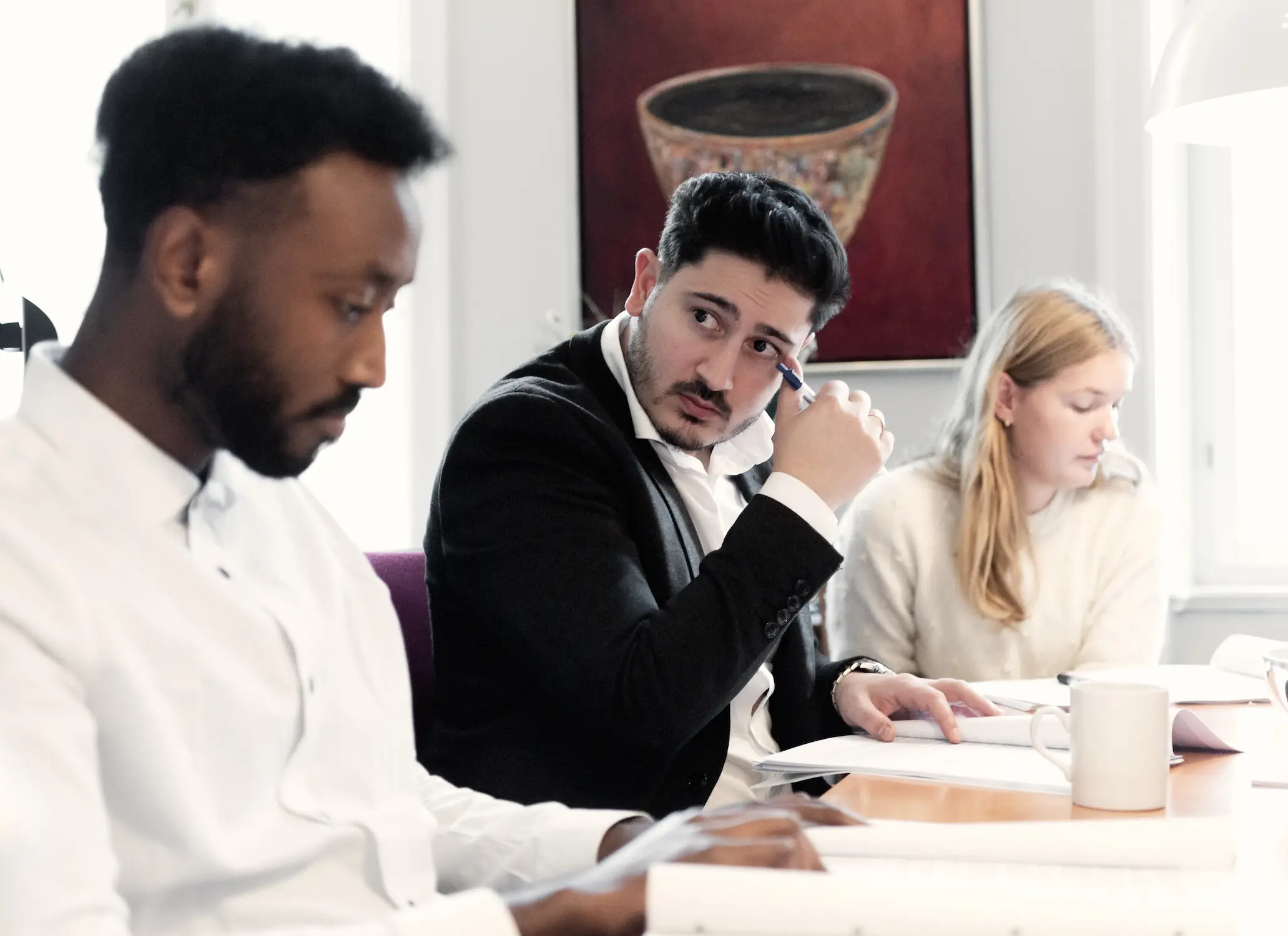 Tre personer sidder ved et mødebord og gennemgår dokumenter. En mand i jakkesæt ser op og taler, mens han holder en pen, og en mand i hvid skjorte læser i papirerne ved siden af. I baggrunden sidder en kvinde og arbejder med sine dokumenter, og der står en kop på bordet.