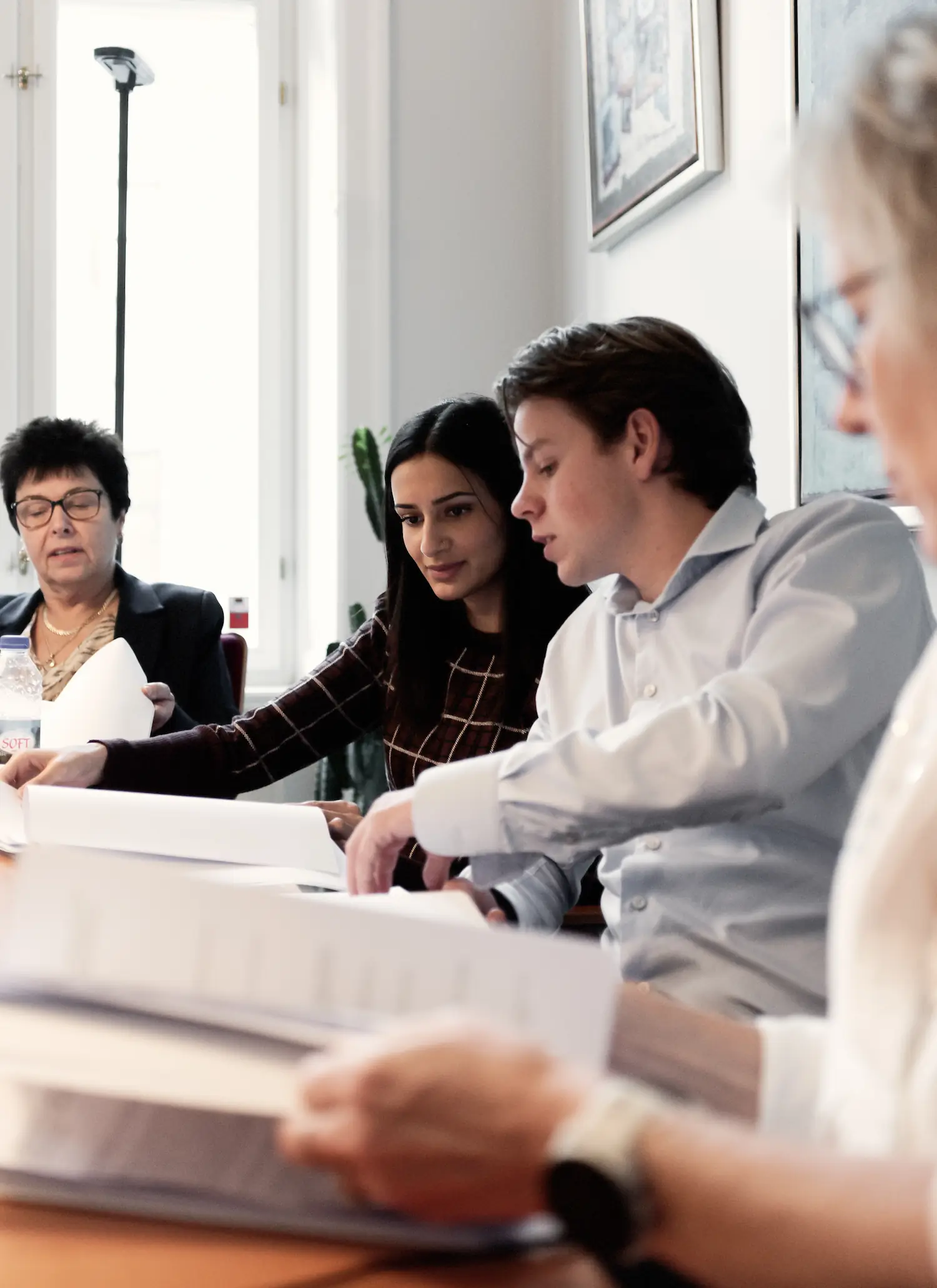 Tre personer sidder ved et mødebord og arbejder med dokumenter. En mand i jakkesæt taler og gestikulerer med en pen, mens en mand i hvid skjorte kigger ned på papirerne. I baggrunden sidder en kvinde og læser, og der står en kop på bordet.