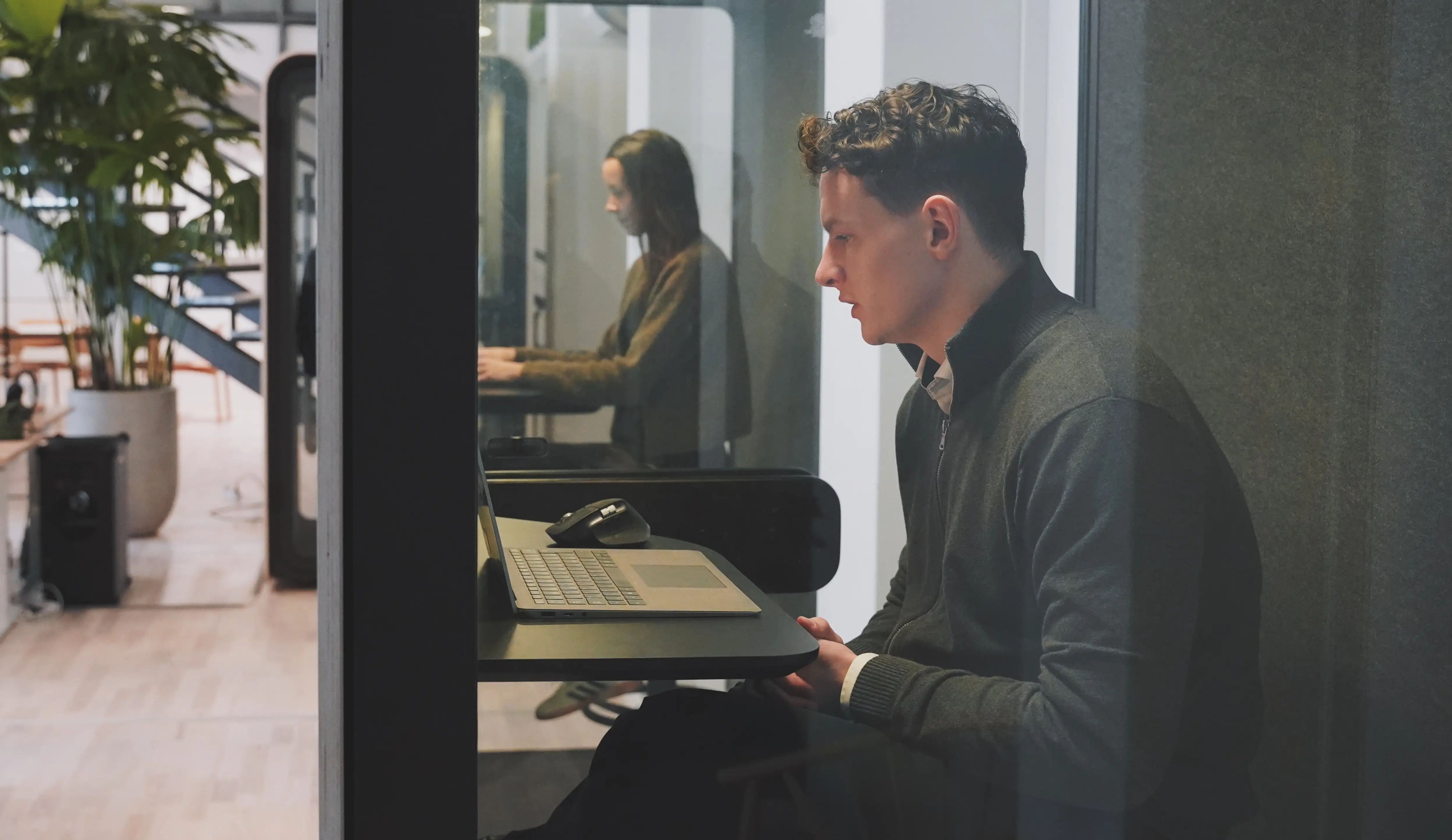 A man working on a laptop inside a glass-walled office pod in a modern workspace.