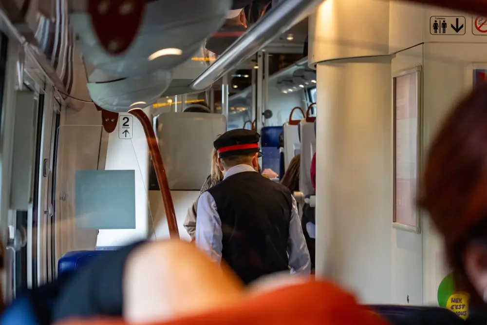 Vue de l'intérieur d'un train, montrant un agent commercial SNCF de dos, portant une casquette et un gilet noir sur une chemise blanche, se dirigeant vers le fond du wagon. On aperçoit d'autres passagers et des éléments de l'intérieur du train.