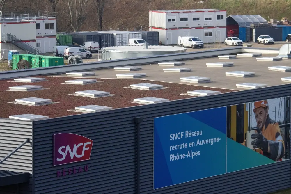 Une vue extérieure d'un bâtiment moderne avec un toit végétalisé, et un grand panneau publicitaire affichant "SNCF Réseau recrute en Auvergne-Rhône-Alpes" avec l'image d'un travailleur SNCF en casque de sécurité. En arrière-plan, on aperçoit des conteneurs modulaires et des véhicules de service.