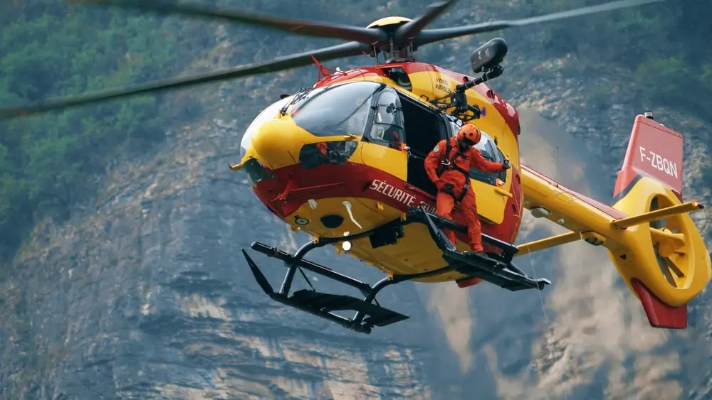 Un hélicoptère de sauvetage jaune et rouge est en vol, avec une personne vêtue d'une combinaison de sauvetage orange se penchant hors de la porte. L'hélicoptère porte l'inscription "SÉCURITÉ CIVILE". En arrière-plan, on distingue une paroi rocheuse ou une montagne.