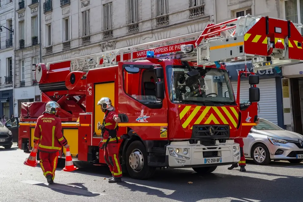 Un camion de pompiers rouge vif des Sapeurs-Pompiers de Paris est stationné dans une rue de ville. Deux pompiers en tenue de protection rouge et jaune se tiennent près du véhicule, l'un d'eux plaçant un cône de signalisation. Le camion est équipé d'une grande échelle déployée. Des bâtiments parisiens classiques sont visibles en arrière-plan.