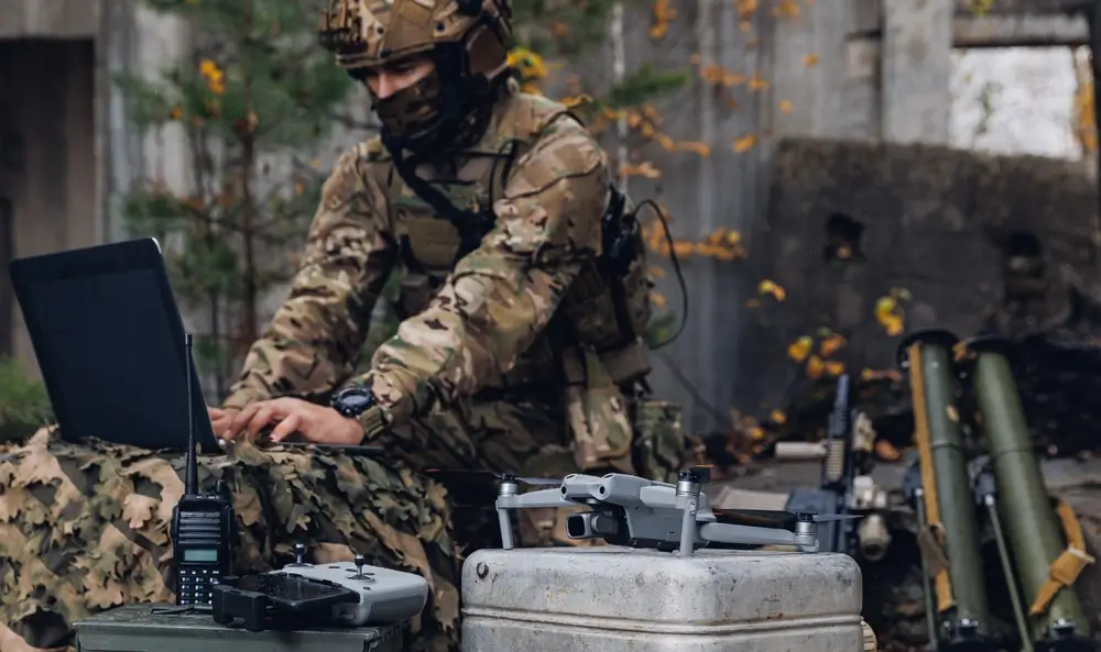 Un soldat en tenue de camouflage et casque est assis, les mains sur un ordinateur portable. Devant lui, un drone et sa télécommande, ainsi qu'une radio. En arrière-plan, on aperçoit d'autres équipements militaires et des débris.